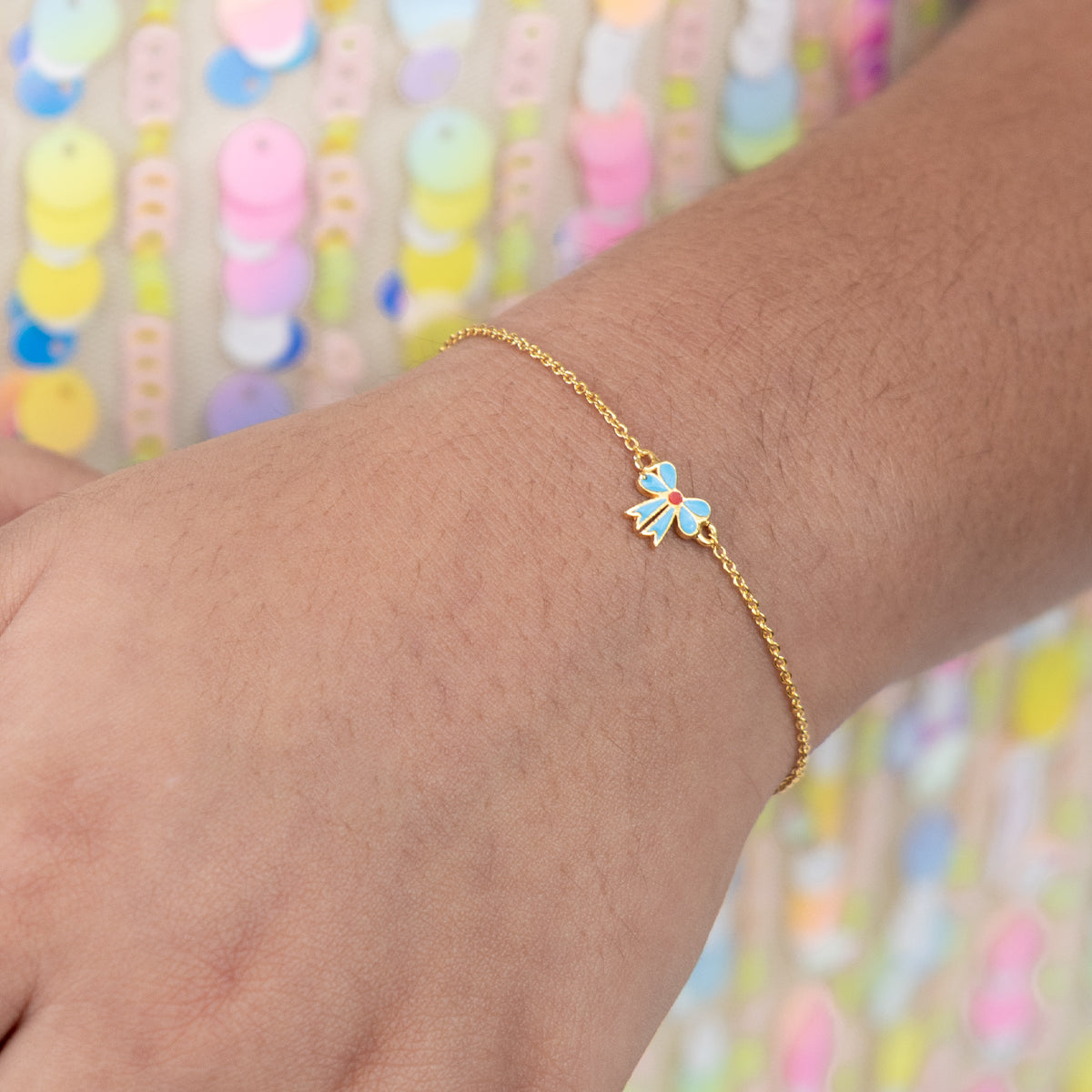 Tiny Jewels gold bracelet with a colorful flower charm on a wrist against a multicolored background