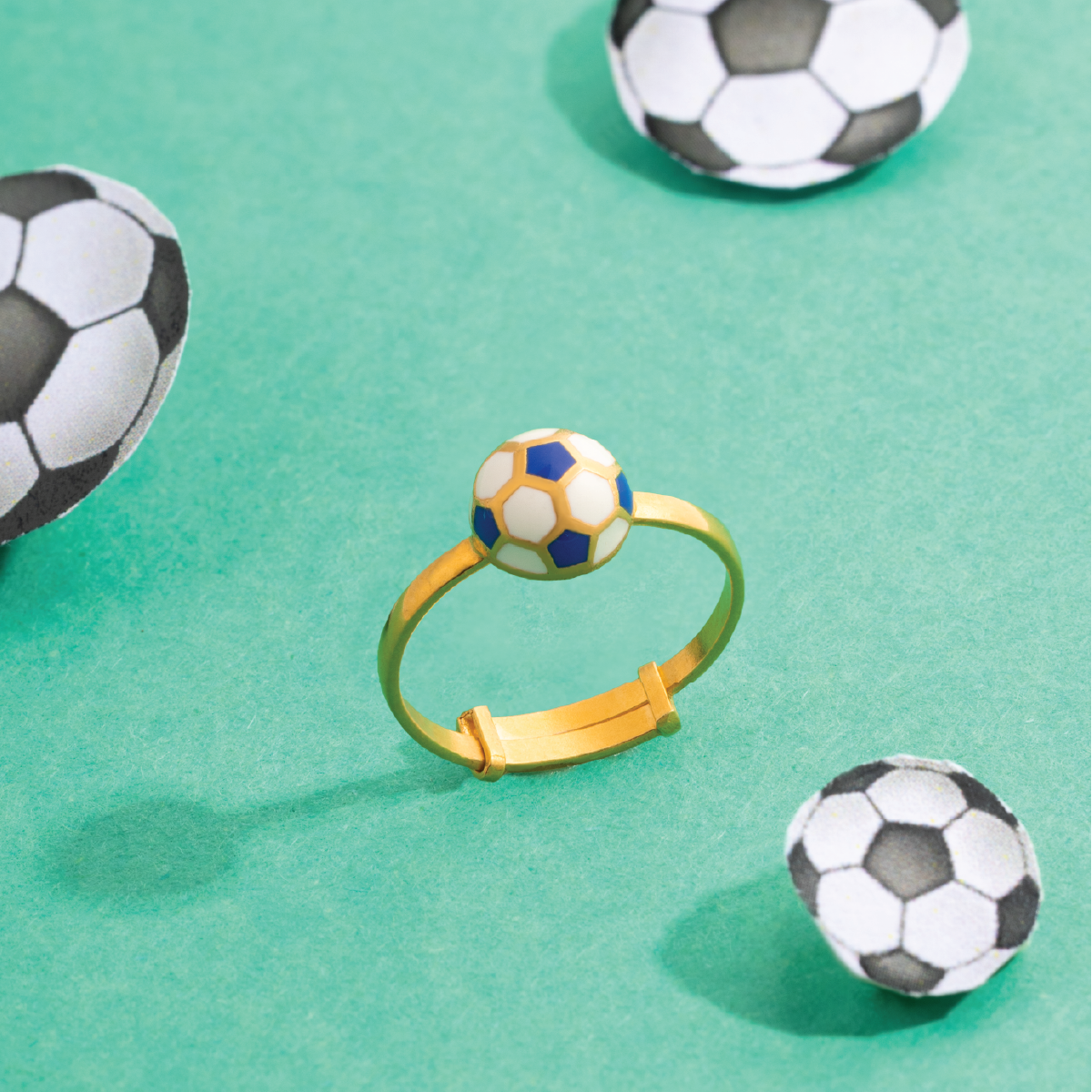 Tiny Jewels gold ring with a soccer ball charm on a green background with soccer ball decorations.
