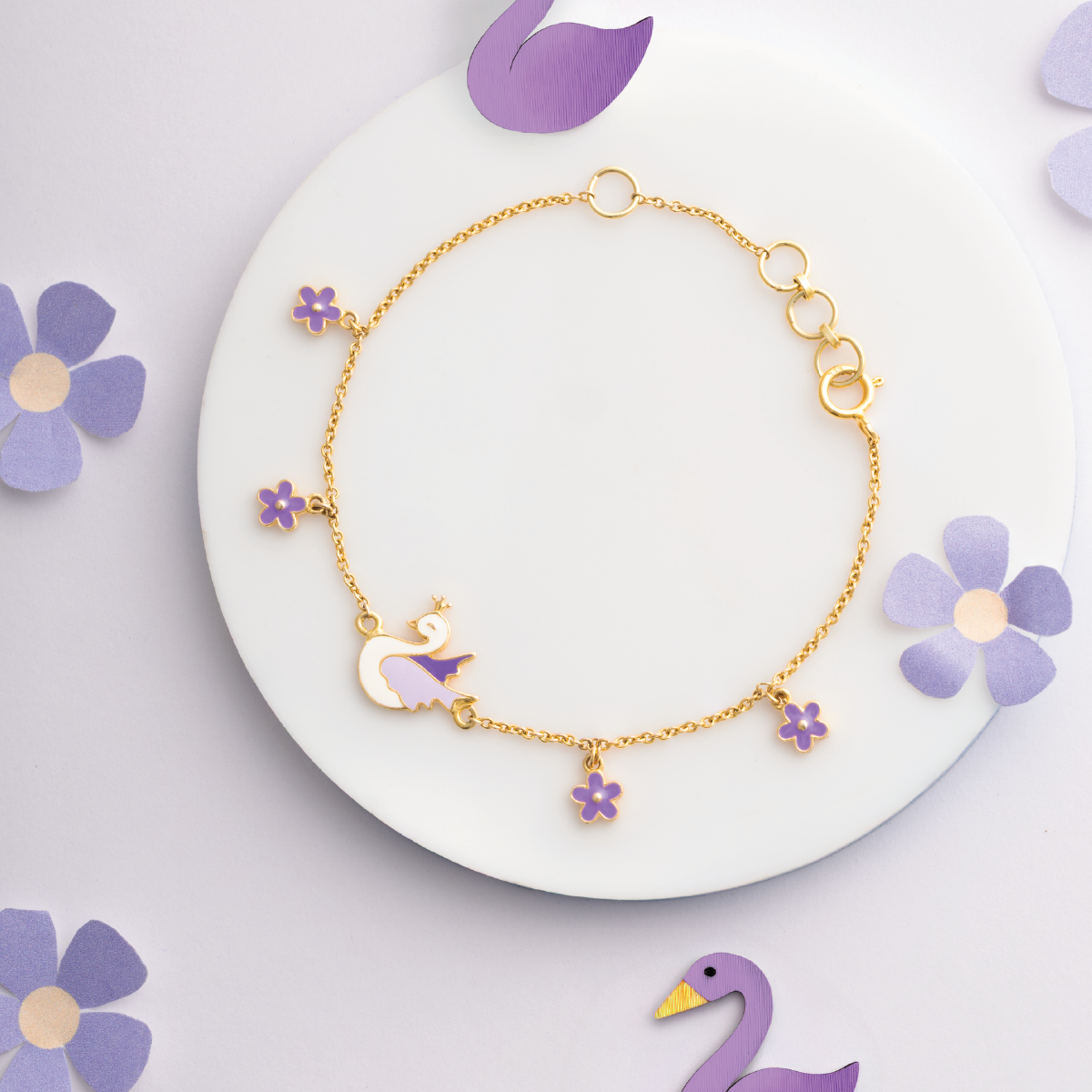 Tiny Jewels gold bracelet with purple swan and flower charms on a white plate with purple flowers.