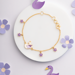 Tiny Jewels gold bracelet with purple swan and flower charms on a white plate with purple flowers.