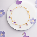 Tiny Jewels gold bracelet with purple swan and flower charms on a white plate with purple flowers.
