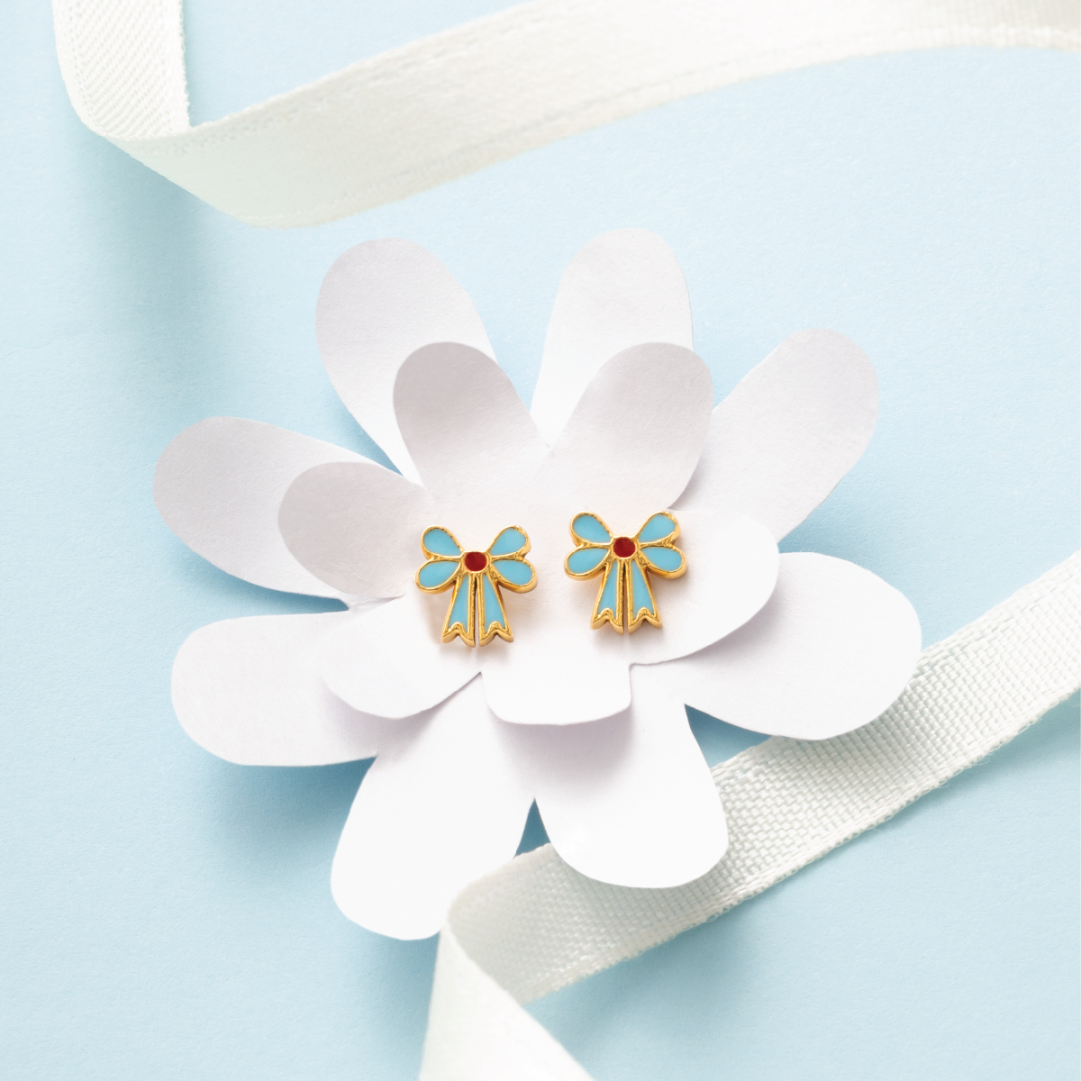 Tiny Jewels gold and blue bow-shaped earrings on a white flower with a light blue background