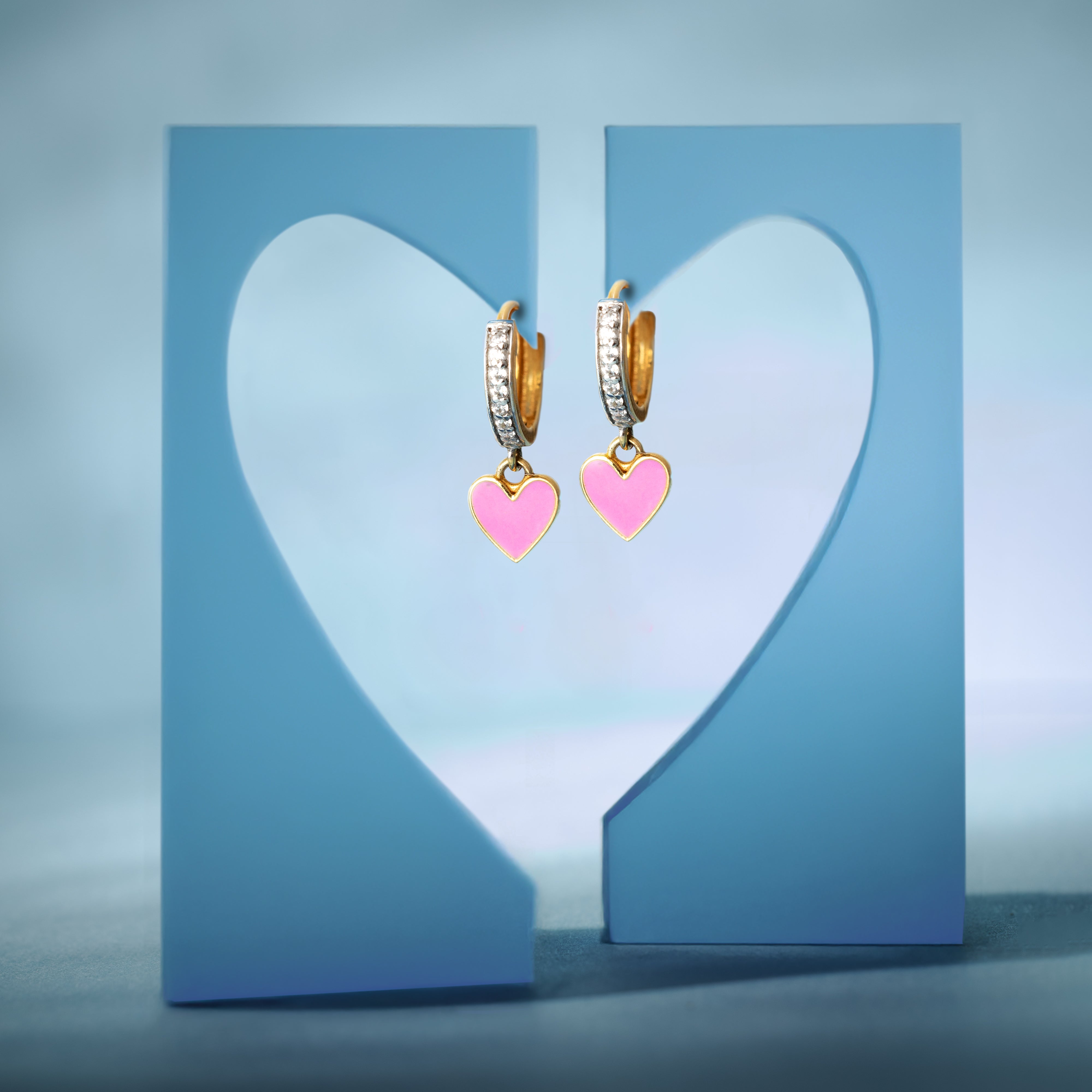 Tiny Jewels pair of gold hoop earrings with pink heart charms on a blue heart-shaped stand.