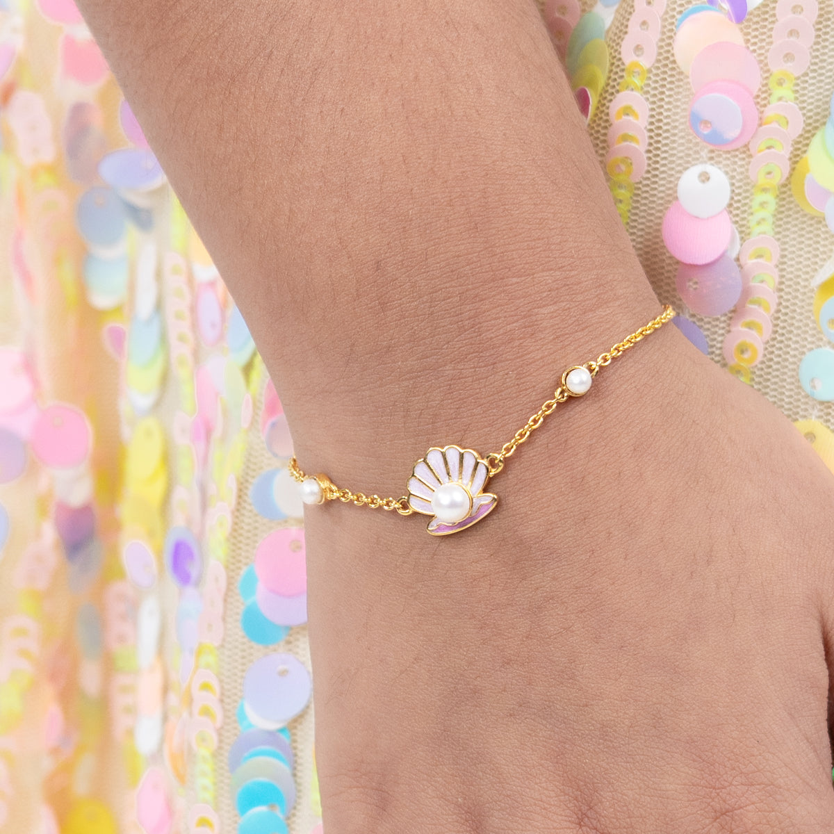 Tiny Jewels gold bracelet with a shell and pearl charm on a wrist against a colorful sequin background