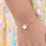 Tiny Jewels gold bracelet with a shell and pearl charm on a wrist against a colorful sequin background