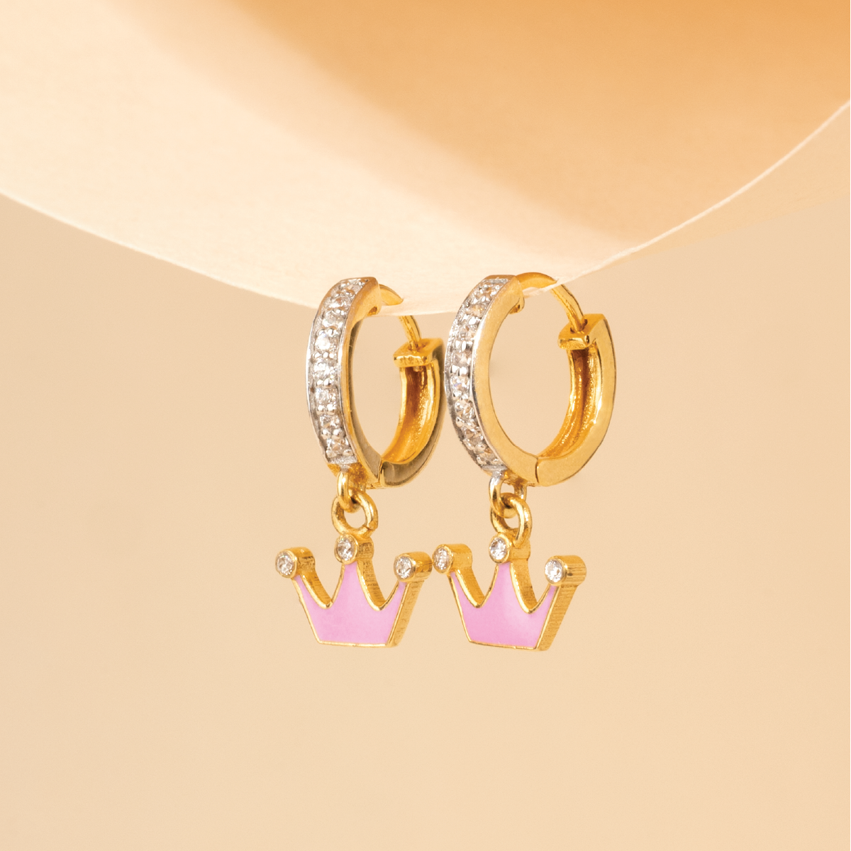 Tiny Jewels gold hoop earrings with pink crown charms on a beige background