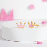 Tiny Jewels pink and gold crown-shaped earrings on a white stand with pink decorative elements in the background.