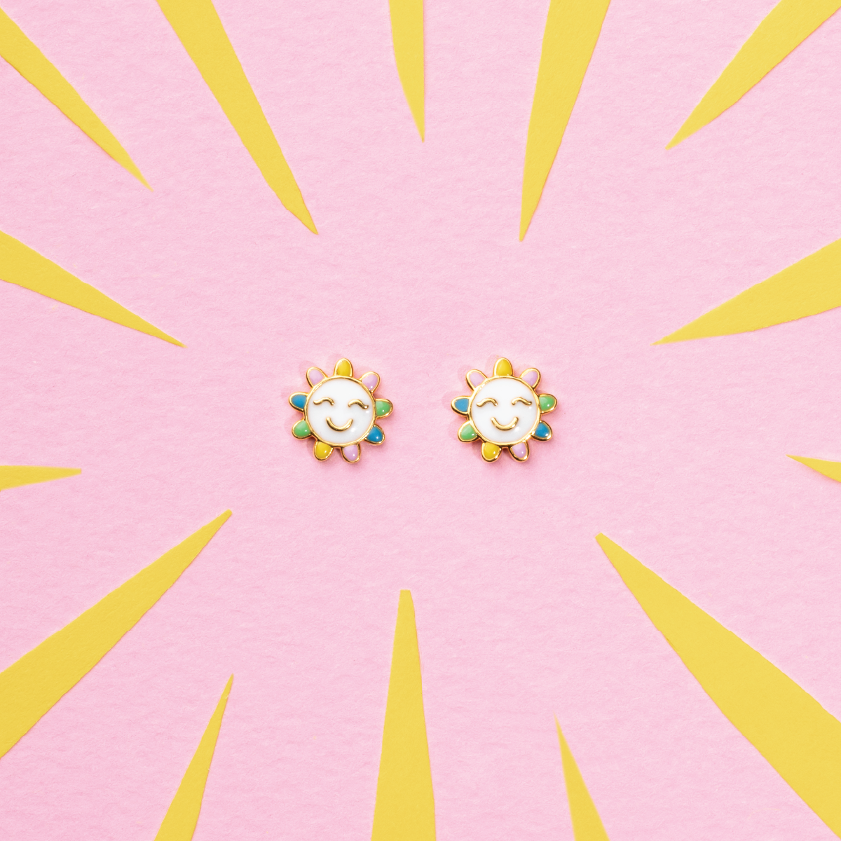 Tiny Jewels sun-shaped earrings on a pink background with yellow sunburst design