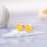 Tiny Jewels gold flower-shaped earrings on a white surface with a blurred background