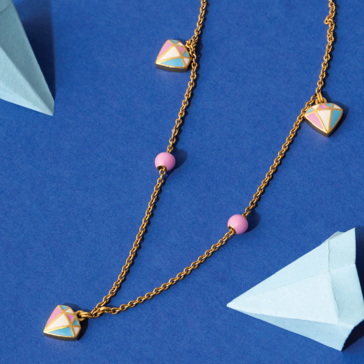 Tiny Jewels necklace with geometric pendants and pink beads on a blue background