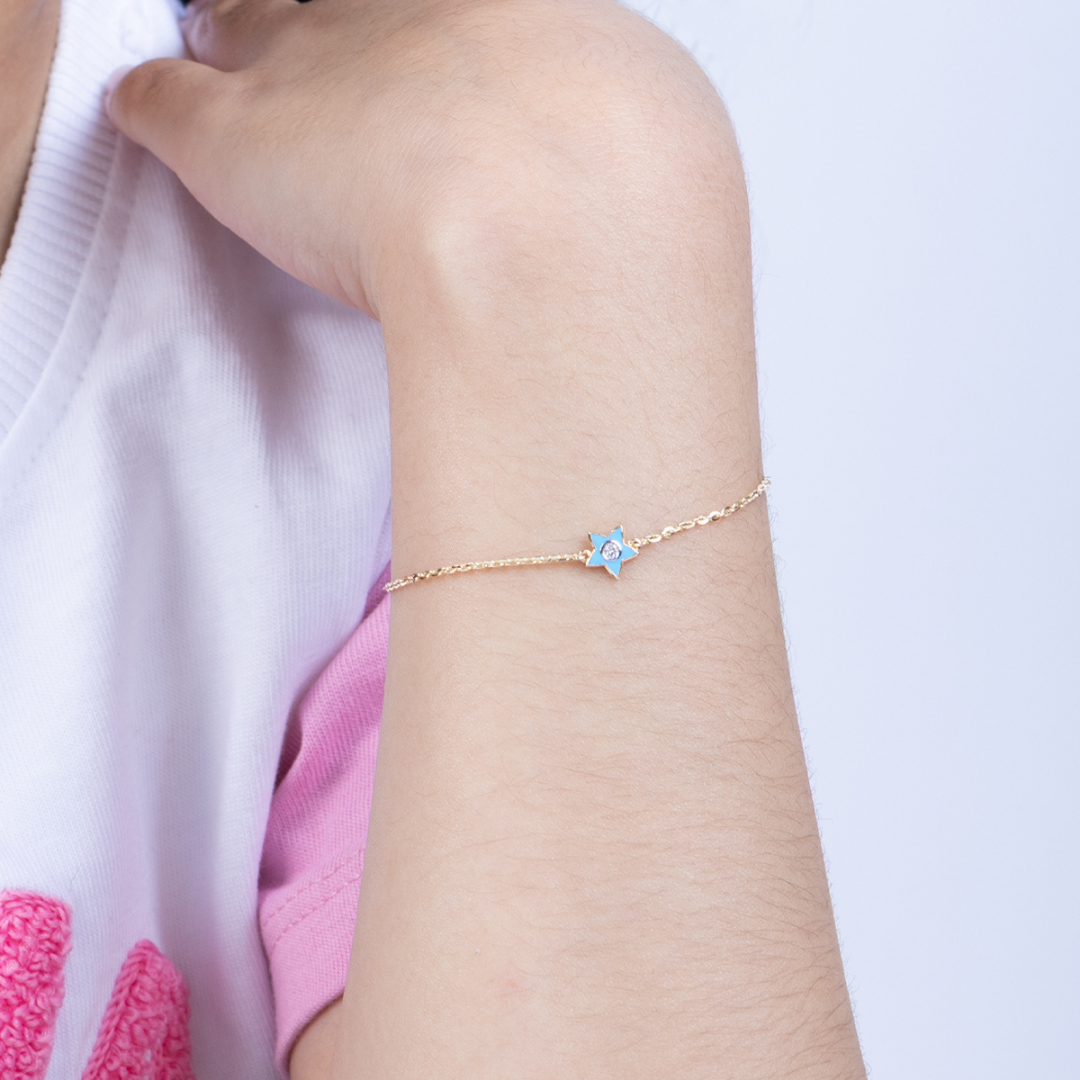Tiny Jewels gold bracelet with a blue star charm on a person's wrist against a light pink background