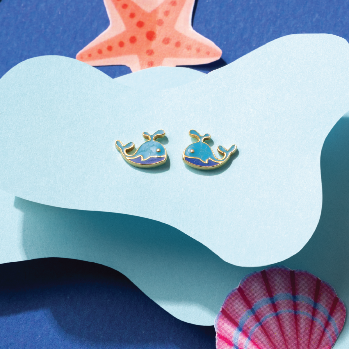 Tiny Jewels pair of whale-shaped earrings on a blue and white wave background with a starfish and shell.