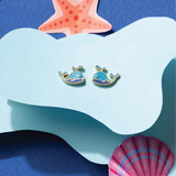 Tiny Jewels pair of whale-shaped earrings on a blue and white wave background with a starfish and shell.